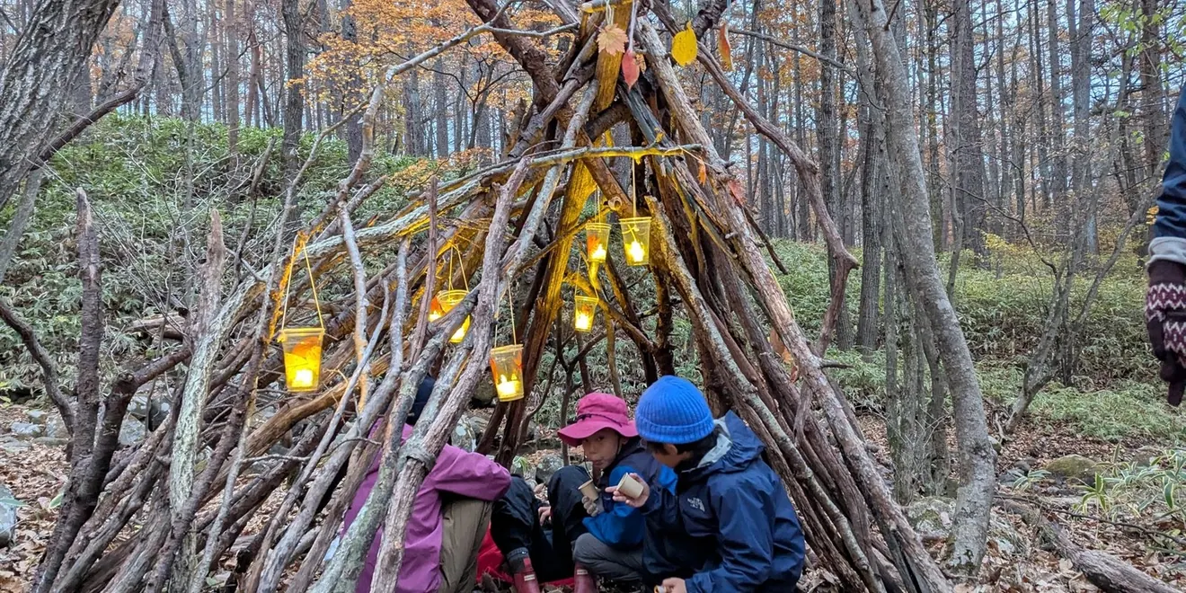 森で枝の小屋とランタンを囲む体験写真（当年公式案内掲載）